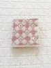 The Organic Cotton Baby Blanket - Bouquet features a folded pink and cream quilted design with a diamond pattern and lace edging, resting on a larger white quilted surface.