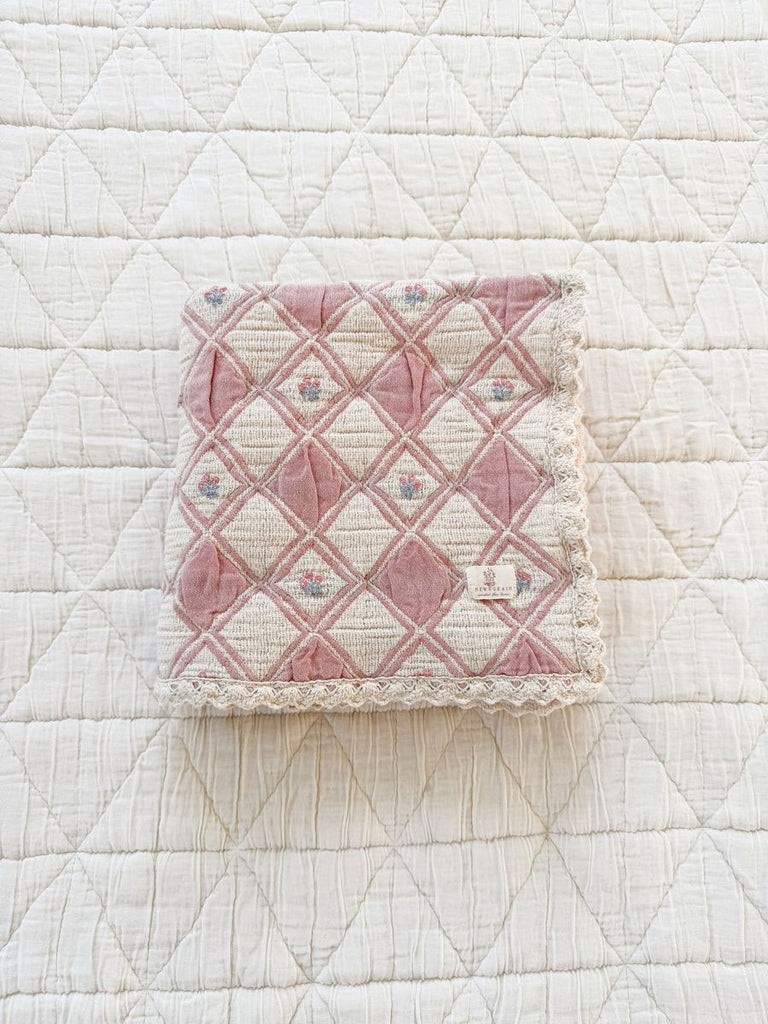 The Organic Cotton Baby Blanket - Bouquet features a folded pink and cream quilted design with a diamond pattern and lace edging, resting on a larger white quilted surface.