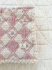 A close-up of the Organic Cotton Baby Blanket - Bouquet, a vintage-style, pink and cream diamond-patterned blanket with lace trim and a "Neverplain" label, crafted from organic cotton in Portugal on a quilted white surface.