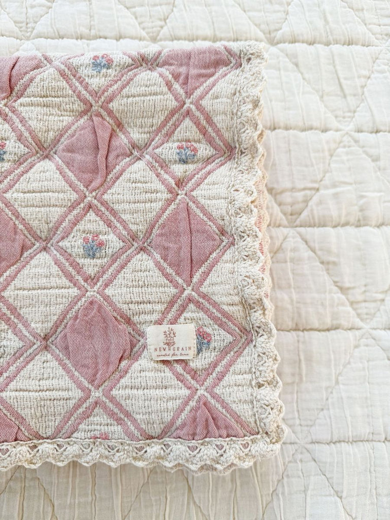 A close-up of the Organic Cotton Baby Blanket - Bouquet, a vintage-style, pink and cream diamond-patterned blanket with lace trim and a "Neverplain" label, crafted from organic cotton in Portugal on a quilted white surface.