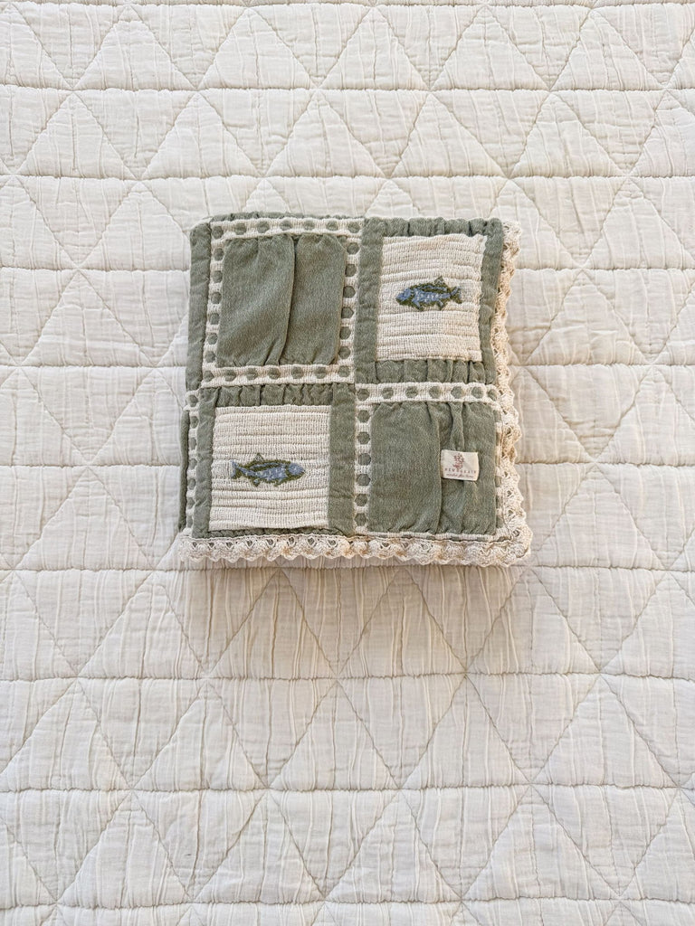The Organic Cotton Baby Blanket - Fishing, with a quilted white diamond pattern, holds a closed cloth book made of green and cream fabric, adorned with embroidered fish and a lace border.