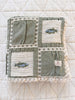 The Organic Cotton Baby Blanket - Fishing, folded to showcase its green fabric with cream crochet trim, embroidered fish, and quilted square patterns, is displayed on a white quilted surface.
