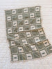 The Organic Cotton Baby Blanket - Fishing features a green and cream grid pattern with small embroidered fish, crafted from organic cotton in Portugal, shown displayed on top of a larger white quilted surface.