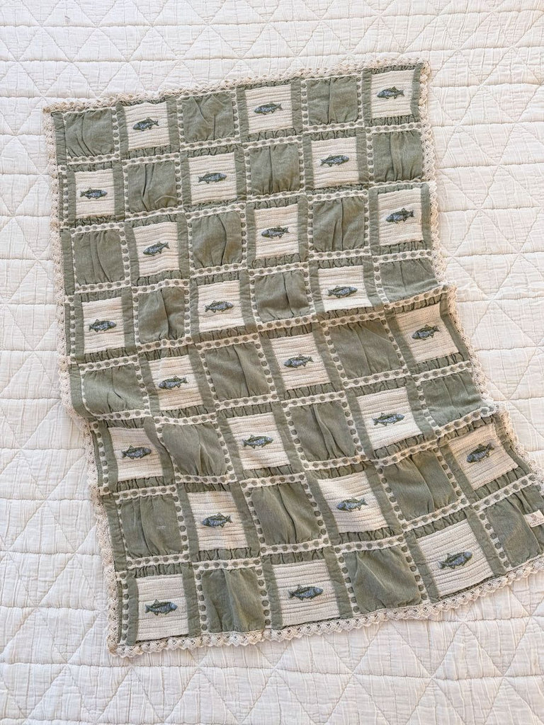 The Organic Cotton Baby Blanket - Fishing features a green and cream grid pattern with small embroidered fish, crafted from organic cotton in Portugal, shown displayed on top of a larger white quilted surface.