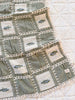 The Organic Cotton Baby Blanket - Fishing features green and beige patchwork squares, some with embroidered fish, crocheted lace edging, and is shown on a white textured background.