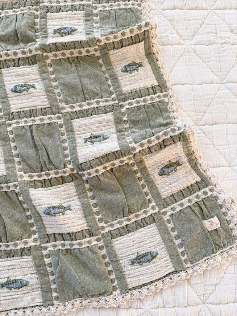 The Organic Cotton Baby Blanket - Fishing features green and beige patchwork squares, some with embroidered fish, crocheted lace edging, and is shown on a white textured background.