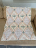The Organic Cotton Quilt - Granny, crafted in Portugal from organic cotton, features a pastel patchwork of pink, cream, and light green with diamond patterns and floral designs. Lace trim accents the edges, and the quilt is elegantly draped over a beige couch.