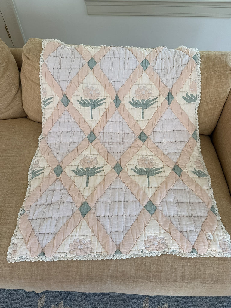 The Organic Cotton Quilt - Granny, crafted in Portugal from organic cotton, features a pastel patchwork of pink, cream, and light green with diamond patterns and floral designs. Lace trim accents the edges, and the quilt is elegantly draped over a beige couch.