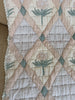 The Organic Cotton Quilt - Granny, made in Portugal from organic cotton, features a diamond pattern adorned with pastel pink, cream, and green floral designs. Delicate lace trim accents the edge of the quilt, which is draped over a beige surface. Ships in one week.