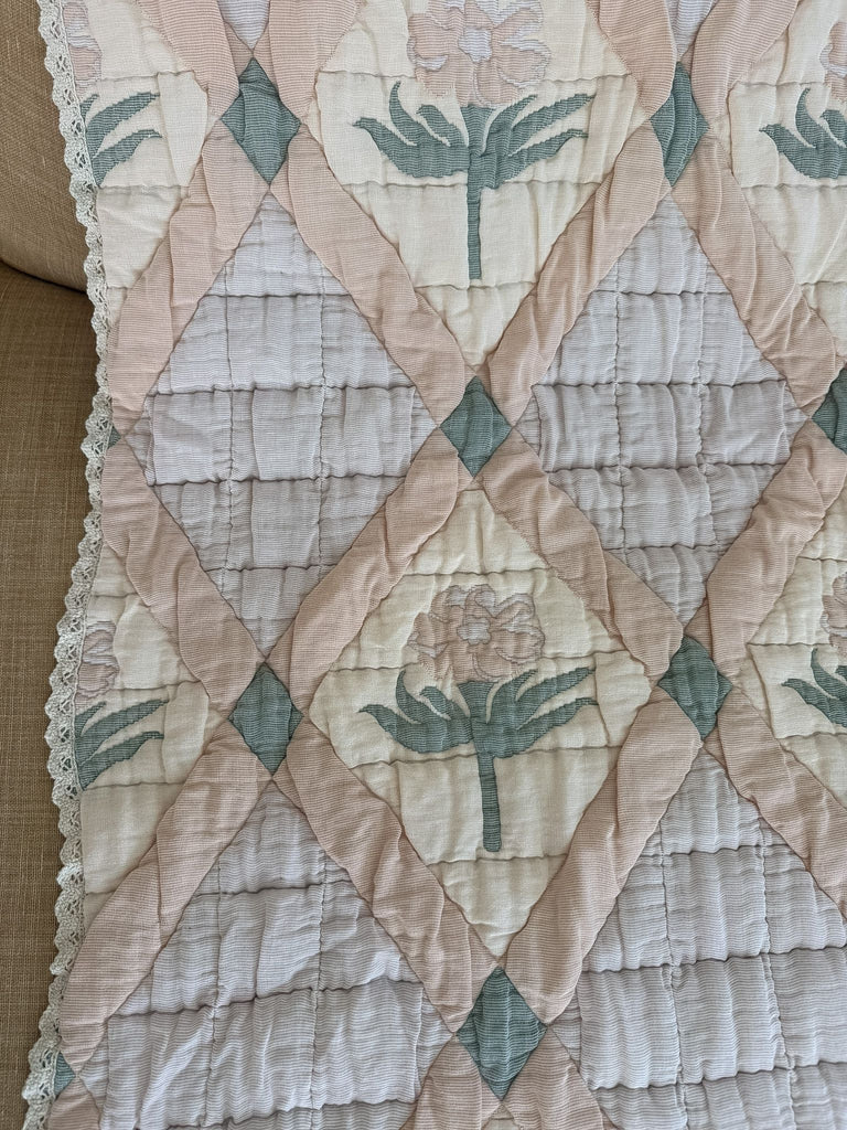 The Organic Cotton Quilt - Granny, made in Portugal from organic cotton, features a diamond pattern adorned with pastel pink, cream, and green floral designs. Delicate lace trim accents the edge of the quilt, which is draped over a beige surface. Ships in one week.