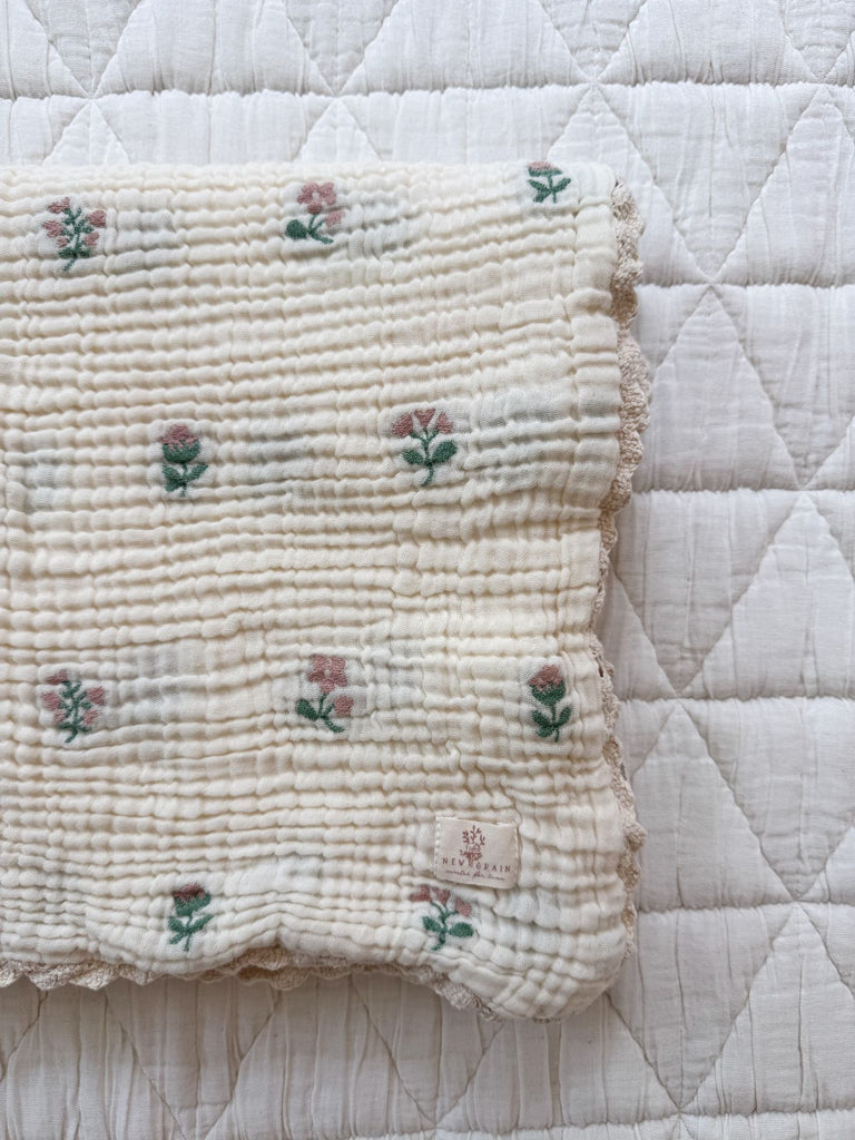 A cream-colored Organic Cotton Layered Gauze Blanket - Dahlia, featuring a quilted design with delicate pink and green floral patterns, scalloped edges, and a small fabric label, is crafted from soft 8-layer organic cotton gauze and displayed on a white quilted surface.