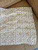 The Organic Cotton Quilt - Daffodil, crafted in Portugal from organic cotton, features a scalloped lace edge and square patches adorned with beige and pink floral patterns in soft pastel colors. Draped over a tan couch, this quilt ships in one week.