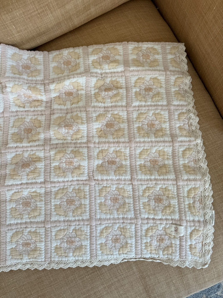 The Organic Cotton Quilt - Daffodil, crafted in Portugal from organic cotton, features a scalloped lace edge and square patches adorned with beige and pink floral patterns in soft pastel colors. Draped over a tan couch, this quilt ships in one week.