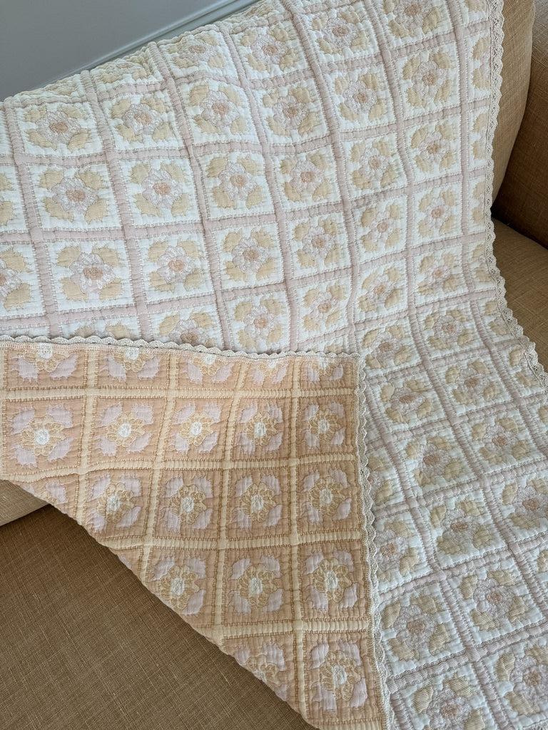 An Organic Cotton Quilt - Daffodil, featuring a cream and pastel pink floral square pattern, is draped over a beige couch; made from organic cotton and ships in one week.