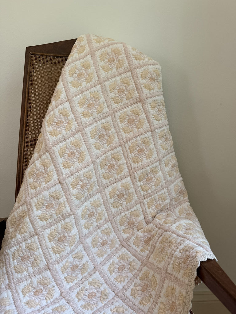 The Organic Cotton Quilt - Daffodil, crafted in Portugal from organic cotton in cream and pale yellow hues with a repeating floral pattern, drapes over a wooden chair with a woven backrest against a plain white wall.