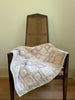 The Organic Cotton Quilt - Sundrop (ships in one week), with its pastel patchwork and floral pattern, drapes over a wooden chair featuring a woven cane back, set against a plain wall and wooden floor.