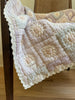 An Organic Cotton Quilt - Sundrop (ships in one week) with a delicate lace edge is draped over a wooden chair. Crafted from organic cotton, the quilt displays a patchwork pattern in soft pink and blue hues. A wooden floor and cream wall are visible in the background.