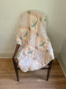 An Organic Cotton Quilt - Granny (ships in one week), hand dyed in soft pastels with a delicate leaf pattern, is draped over a wooden chair with a woven seat, resting on a hardwood floor against a white wall. Crafted from organic cotton and made in Portugal, this quilt adds natural charm and timeless elegance to any space.