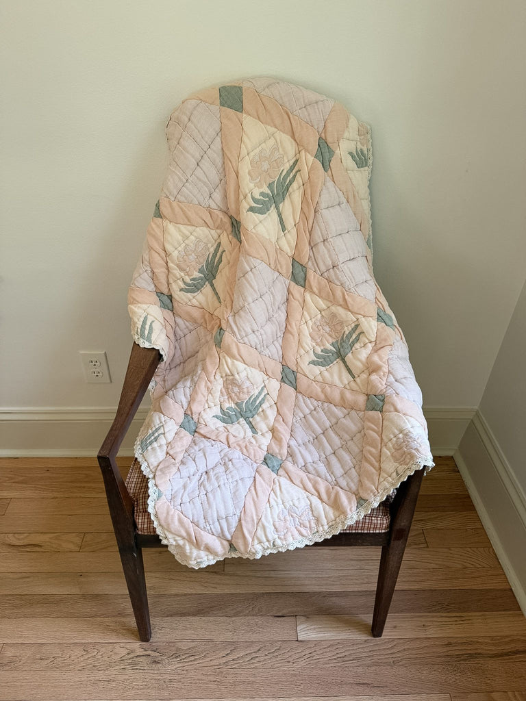 An Organic Cotton Quilt - Granny (ships in one week), hand dyed in soft pastels with a delicate leaf pattern, is draped over a wooden chair with a woven seat, resting on a hardwood floor against a white wall. Crafted from organic cotton and made in Portugal, this quilt adds natural charm and timeless elegance to any space.