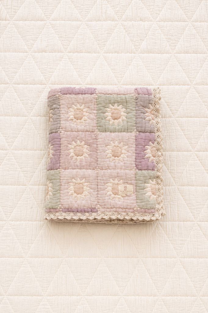 The Organic Cotton Quilt - Sundrop, crafted in Portugal from organic cotton and featuring a pastel patchwork design with floral patterns and delicate lace trim, is displayed on a white, quilted background.