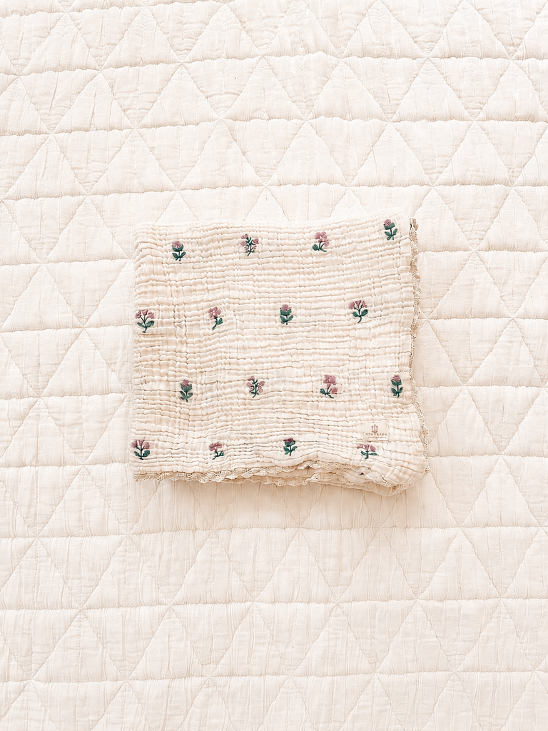 A folded Organic Cotton Layered Gauze Blanket – Dahlia, crafted from soft organic cotton and featuring delicate dahlia embroidery, rests atop a larger diamond-patterned quilted blanket.