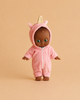 The Minikane Mini Pancha Doll - Prunelle with Unicorn Jumpsuit features a dark-skinned doll dressed in a pink hooded unicorn onesie complete with a golden horn, standing against a beige background and inviting imaginative play. Crafted from high-quality vinyl, this adorable doll inspires creativity and storytelling at every moment.