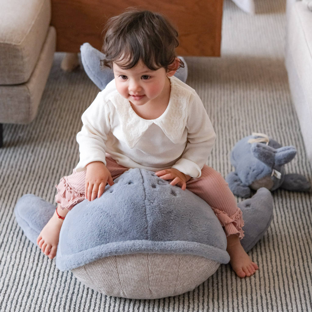A toddler with short brown hair smiles while sitting on a large, plush Ride-On Whale Pouf on a striped carpet. A smaller stuffed animal rests nearby on the floor.