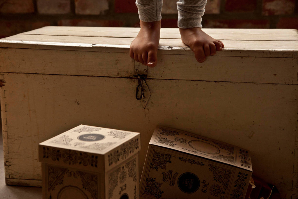 A child’s bare feet stand on a vintage wooden chest near two ornate boxes and FSC certified Natural Square Pyramid Stacker blocks. The warmly lit scene features a brick wall background.