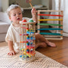 A young child with light brown hair is sitting on a patterned rug playing with the Pound A Ball Tower, using a hammer to hit the top balls. In the background, there's another wooden toy featuring horizontal rainbow tracks in various colors, all made from non-toxic materials.