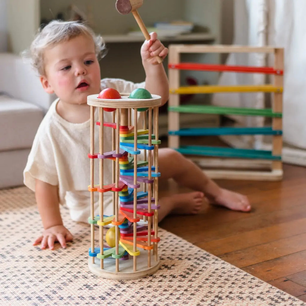 A young child with light brown hair is sitting on a patterned rug playing with the Pound A Ball Tower, using a hammer to hit the top balls. In the background, there's another wooden toy featuring horizontal rainbow tracks in various colors, all made from non-toxic materials.