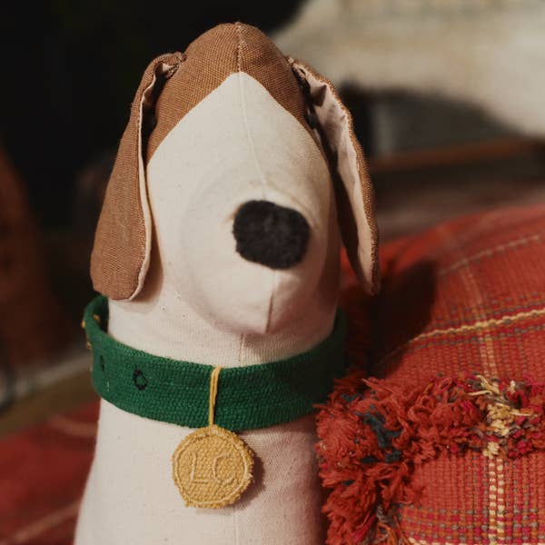 The Dog Cushion - Cooper, featuring floppy brown ears, a black nose, and a green collar with a yellow tag, sits beside a red textured cushion to bring handcrafted charm to your decor.