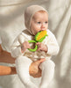 A baby in a cream knit outfit and bonnet sits on an adult’s lap, chewing on the Corn Rattle Toy Rattle. The softly lit, neutral background adds to the cozy scene.