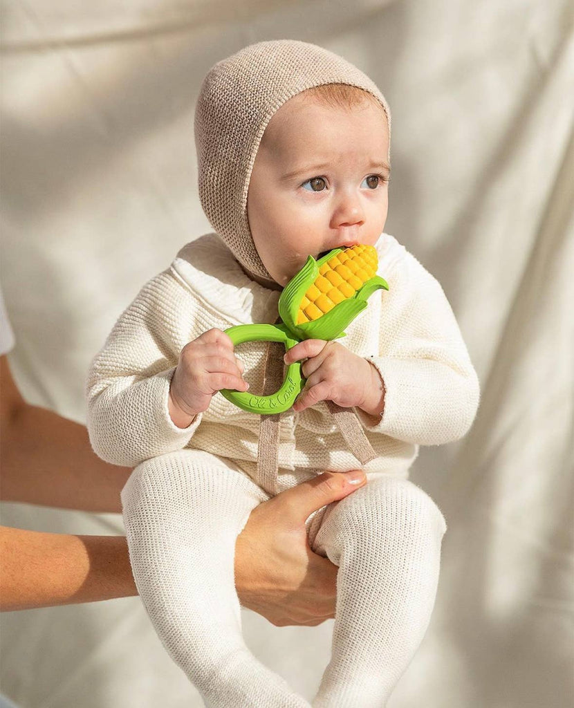 A baby in a cream knit outfit and bonnet sits on an adult’s lap, chewing on the Corn Rattle Toy Rattle. The softly lit, neutral background adds to the cozy scene.
