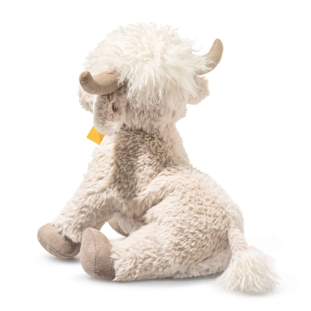 The Steiff Harper Highland Cow is a cream-colored plush nursery toy with shaggy hair, beige horns, a fluffy tail, and brown hooves, shown sitting on a white background.