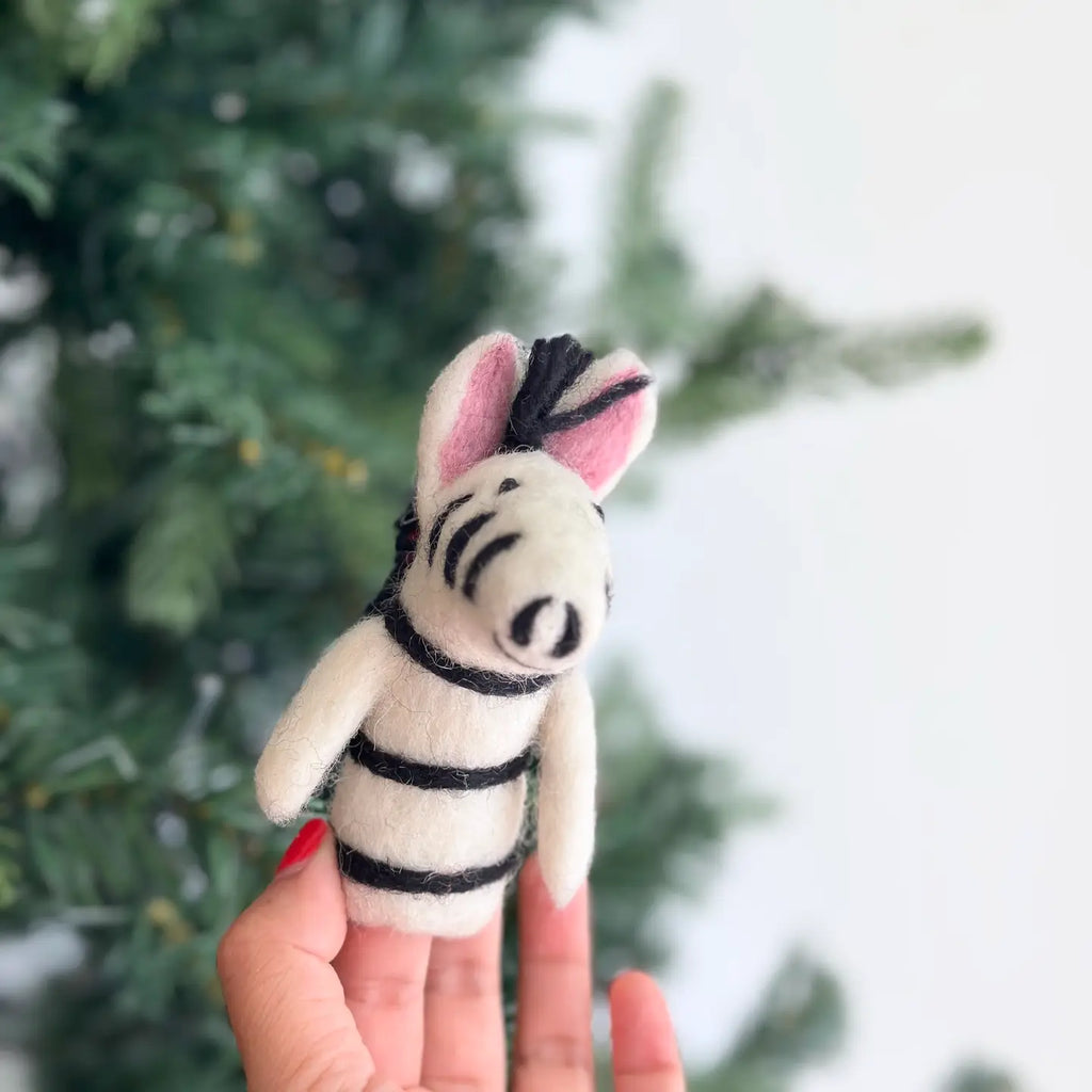 A person with red-painted nails holds a small Zebra Finger Puppet, featuring pink ears and black stripes. The background is blurred but seems to be a green fir tree, making this charming puppet an ideal gift for wild animal lovers.