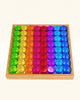 A wooden tray filled with a Bauspiel Lucent Cylinders, 64-Piece Set grid of sixty-four translucent, colored cylinders arranged in columns of green, yellow, orange, pink, red, purple, and blue.