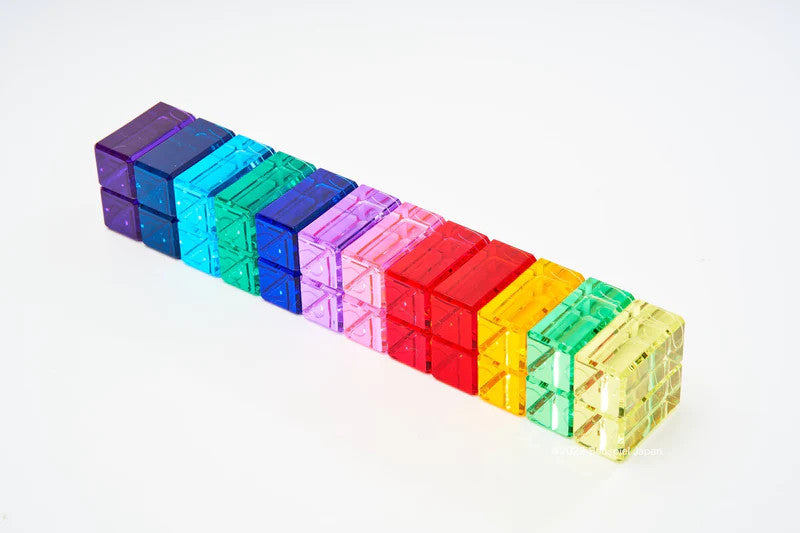 The Bauspiel Luminous Blocks - 24 Piece set features colorful translucent cubes arranged in a gradient, smoothly transitioning from dark purple to light yellow, creating an appealing spectrum against a white background.