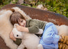 A brown-haired child in a green sweater and blue jeans smiles and hugs the Senger Naturwelt Cuddly Animal - Linen Bear while lying on a furry blanket in a rusty wheelbarrow, surrounded by green grass.