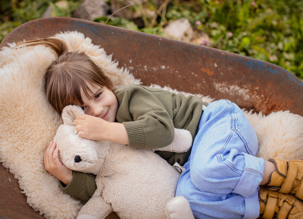 A brown-haired child in a green sweater and blue jeans smiles and hugs the Senger Naturwelt Cuddly Animal - Linen Bear while lying on a furry blanket in a rusty wheelbarrow, surrounded by green grass.