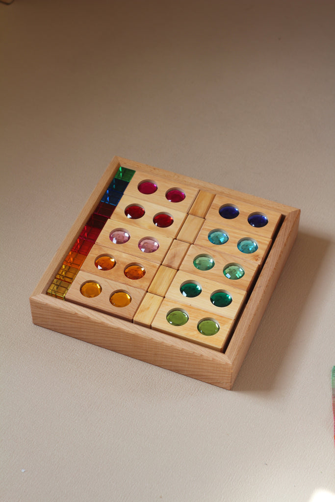 Bauspiel Gem Blocks with Lucent Cubes, 45-Piece Set, displayed as a wooden tray filled with wooden blocks featuring inlaid translucent colorful gems, alongside a stack of small transparent colored cubes.