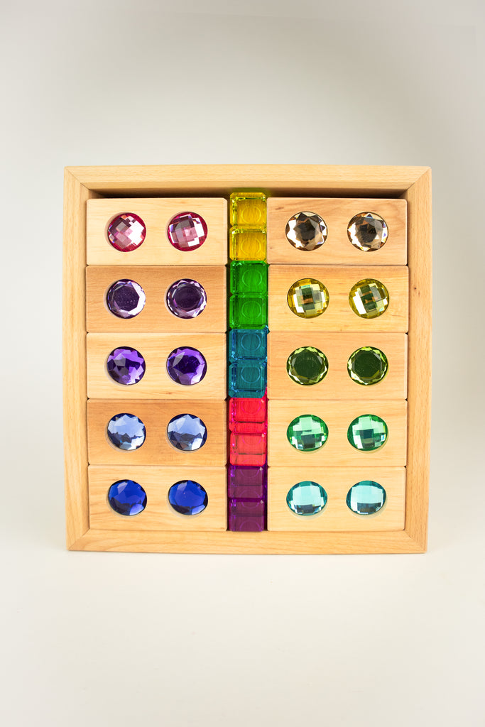 Wooden tray containing ten Bauspiel Gem Blocks with Pastel Lucent Cubes featuring circular colorful gems and a central column of six translucent square cubes.