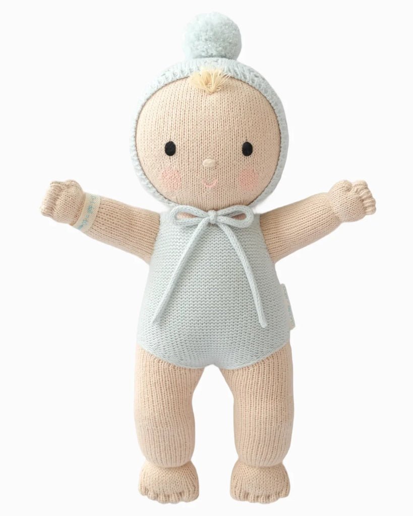 Cuddle + Kind My First Baby (Bluebell) is a soft, hand-knit doll with light skin, outstretched arms, embroidered features, and wears a pale blue romper and pom-pom bonnet. Ships in one week.