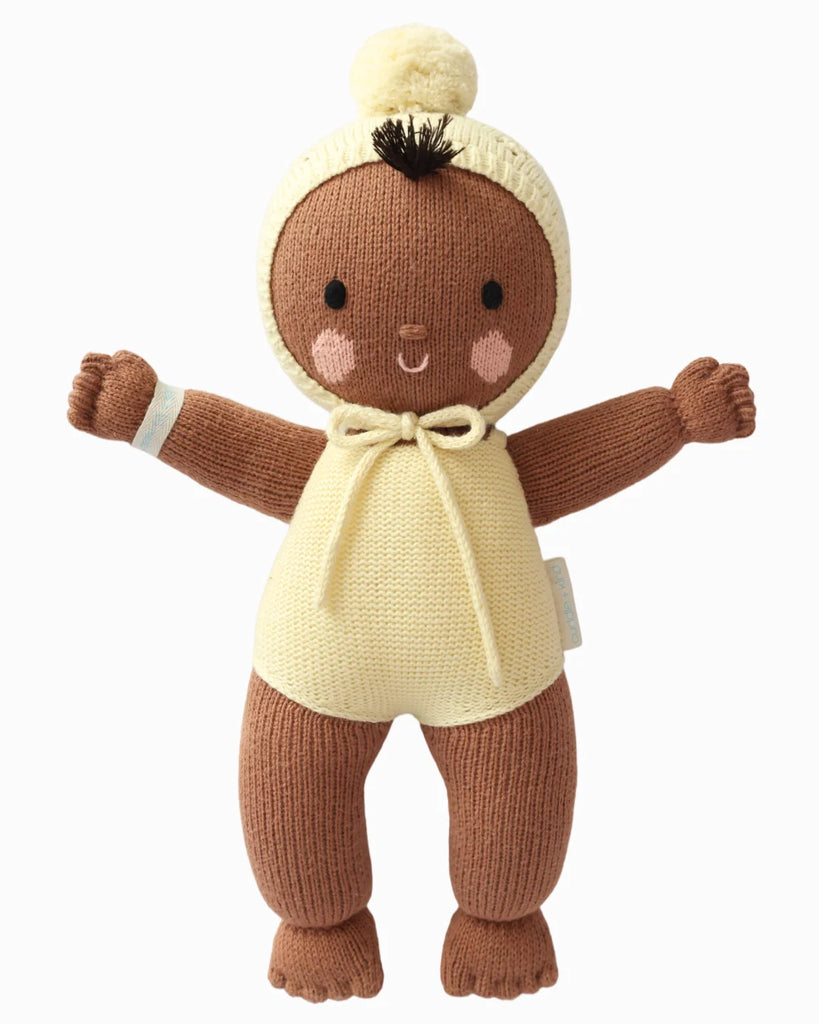 Cuddle + Kind My First Baby (Buttercup) is a hand-knit brown baby doll made from natural cotton yarn, dressed in a cream romper with hood and pom-pom, rosy cheeks, and open arms. Ships in one week.