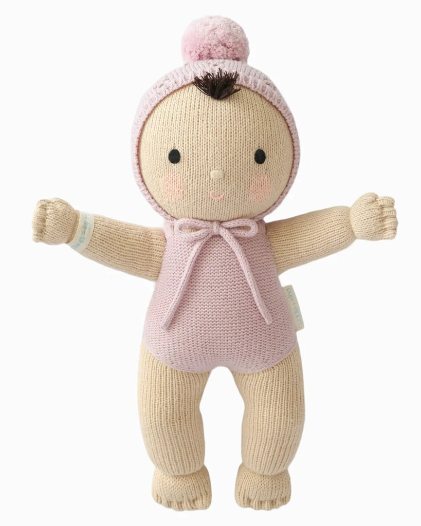 Cuddle + Kind My First Baby (Lavender) is a soft, hand-knit doll made of natural cotton yarn with beige skin, lavender outfit and pom-pom hat, stitched rosy cheeks, small smile, dark brown hair tuft, and outstretched arms. Ships in one week.