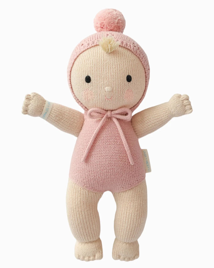 The Cuddle + Kind My First Baby (Rose) doll is hand-knit from natural cotton yarn, wears a pink romper with a matching pom-pom hat, and features embroidered eyes and a smiling face. Fair trade; ships in one week.