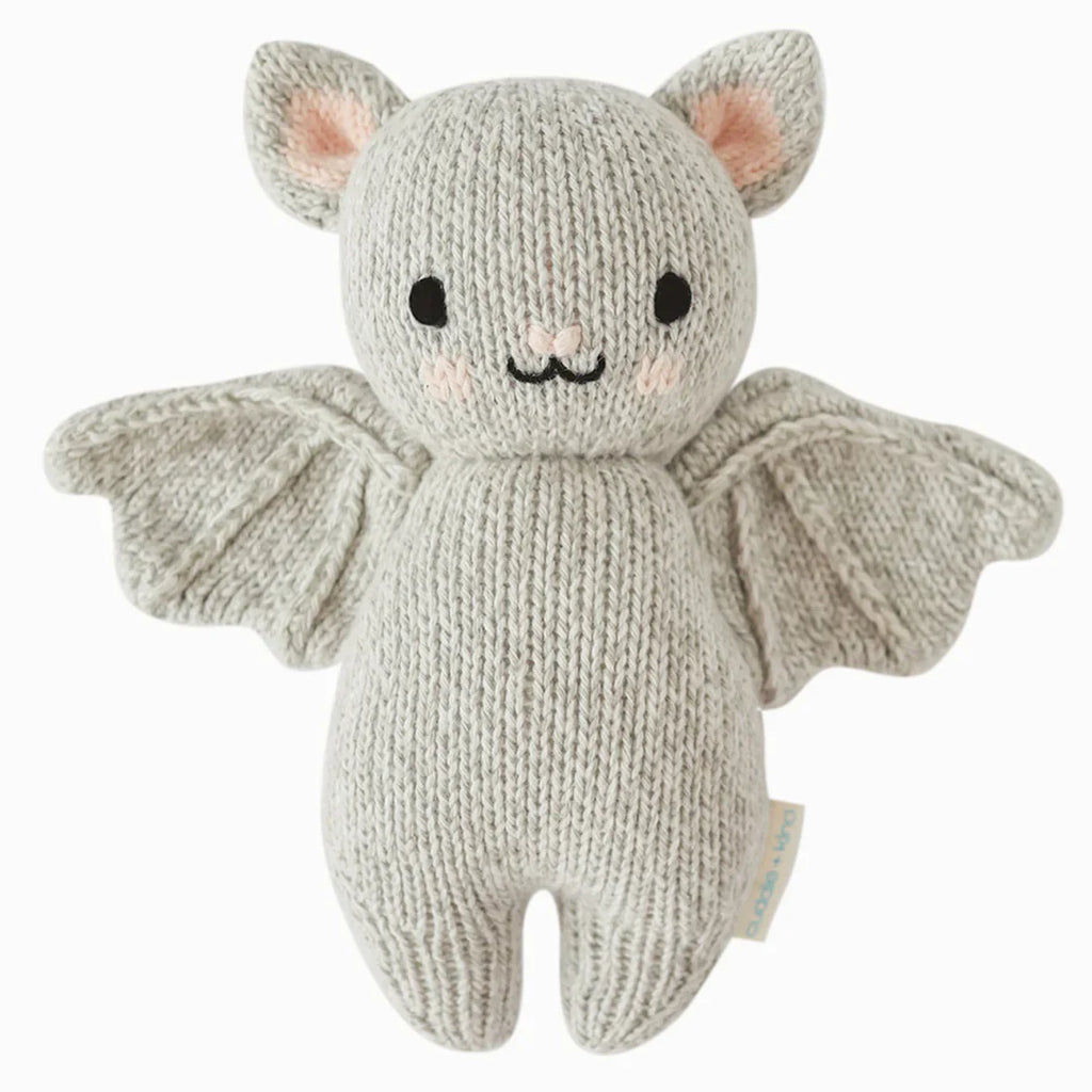 The Cuddle + Kind Baby Limited Edition Grey Bat is a knitted plush toy in the shape of a bat, crafted from light gray Peruvian cotton yarn. It features pink ears, black stitched eyes and mouth, and spread-open wings. A small tag is attached to its side. This hand-knit collectible stands out against the plain white background.