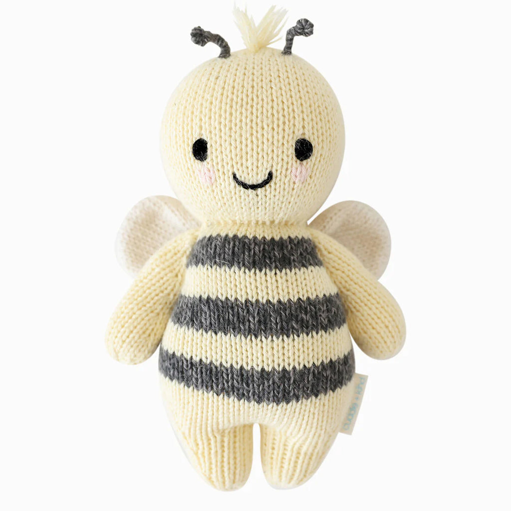 A soft, hand-knit Cuddle + Kind Baby Bee plush toy resembling a bee with black and white stripes, tiny wings, and a smiling face, crafted from Peruvian cotton yarn, isolated on a white background.