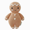 The Cuddle + Kind Baby Gingerbread doll is an heirloom-quality knitted toy in a beige hue, featuring a smiling face crafted from natural Peruvian cotton yarn. It boasts white zigzag patterns on its head and three white buttons, with charming white accents on its feet and hands.