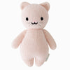 A pink hand-knit stuffed animal resembling a Cuddle + Kind Baby Kitten with a simplified, cute face featuring black dot eyes and a small pink nose, standing against a white background.