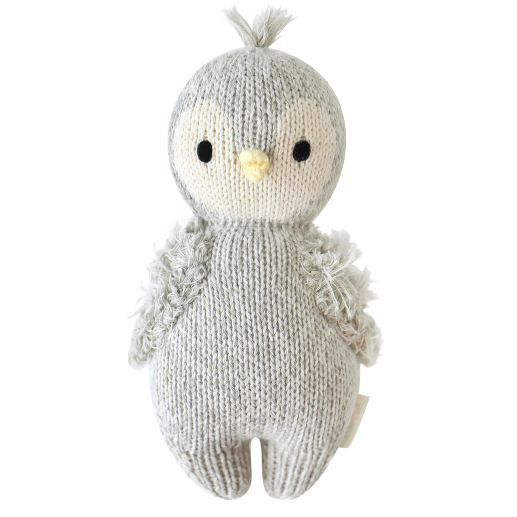 A hand-knit Cuddle + Kind Baby Penguin with a tuft on its head, wing details, and yellow beak, crafted from Peruvian cotton yarn, isolated on a white background.